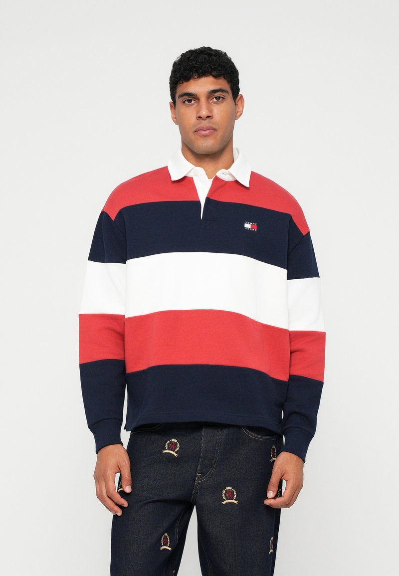 Poloshirt met lange mouwen in rood, wit en marineblauw met horizontale strepen. Heeft een witte kraag en een klein logo op de borst. Draagt bij broeken met borduursel.