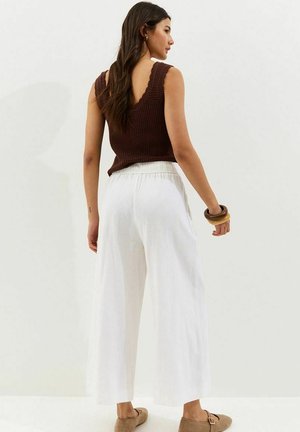 Donna con top marrone senza maniche in maglia, pantaloni bianchi a gamba larga, scarpe slip-on color tan e braccialetti spessi, in piedi davanti a uno sfondo semplice.