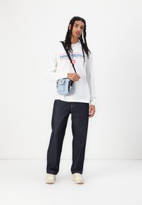 Lys blå crossover-bag med sorte detaljer, båret over en hvit langermet skjorte med rødt og blått logo, kombinert med mørke denim bukser.