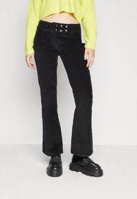 Pantalons en velours côtelé noirs évasés avec une surface texturée et une taille à quatre boutons. Associés à des chaussures noires massives et un pull vert clair vif.