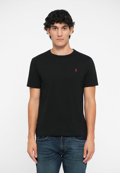 T-shirt en coton noir à col rond, manches courtes, avec un petit logo rouge sur la poitrine gauche. Porté avec un jean en denim bleu.