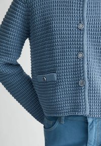 Cardigan lavorato a maglia blu con un motivo testurizzato, dotato di cinque bottoni e una piccola tasca con un accento a bottone. Abbinato a pantaloni blu.