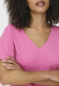 Lyserød bluse med V-hals lavet af struktureret stof, med korte ærmer. Modellens arme er korslagt, hvilket fremviser en glat hudtone.