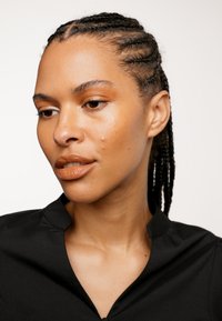 Zwarte stoffen top met een uitsneden kraag, kort geknipte haren met vlechten en een neutrale lipkleur; gladde huid met minimale vlekken.