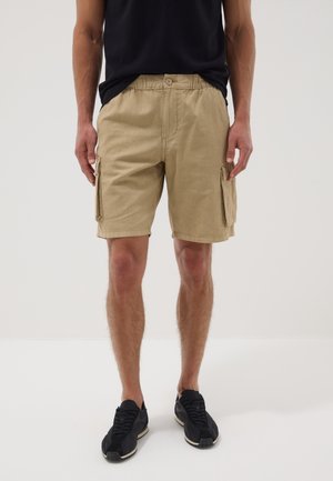 Mann trägt beige Cargoshorts, schwarze Sneaker und ein schwarzes T-Shirt, steht vor einem schlichten hellen Hintergrund.