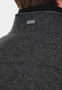 Szary, strukturalny żakiet z subtelnym wzorem w jodełkę; posiada czarny kołnierz oraz metalową metkę z logo marki na plecach.