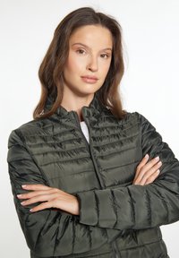 Jachetă de tip puffer verde închis, cu închidere prin fermoar, având tipar de cusături orizontale și un guler înalt. Material neted și lucios, cu o croială confortabilă.