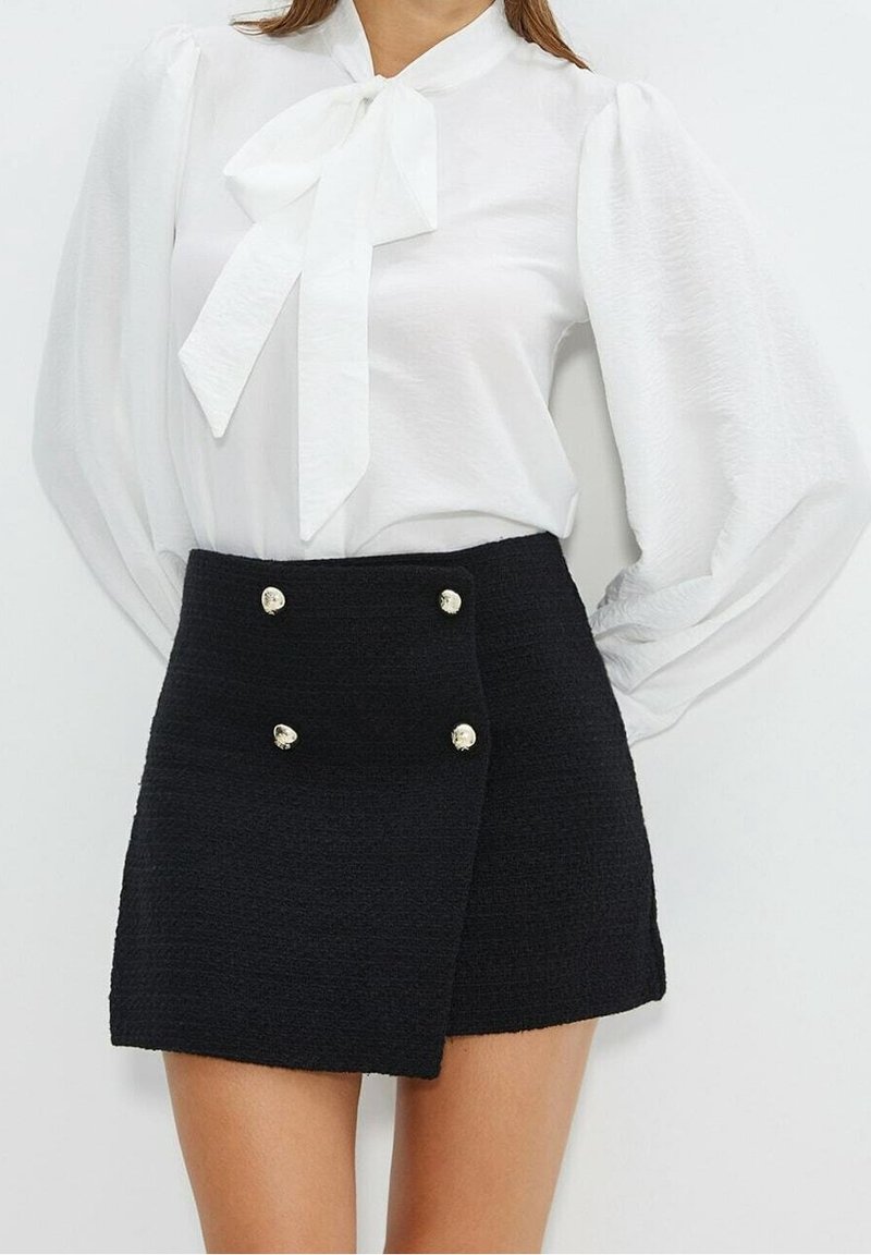 Femme portant un chemisier blanc à manches longues avec un nœud et une mini-jupe noire boutonnée.