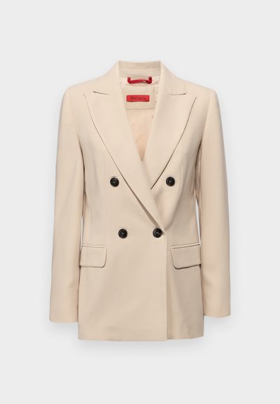 Blazer beige à double boutonnage avec un col cranté, des boutons noirs et deux poches avant. Présente un design ajusté et une texture lisse.
