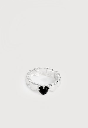 Anello d'argento con bordi smerlati e una pietra nera a forma di cuore al centro su uno sfondo bianco semplice.