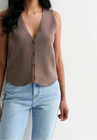 Brun stickad väst med djup V-ringning och femknappsstängning, med en vågig nederkant och tunna axelband, matchad med ljusblåjeans.