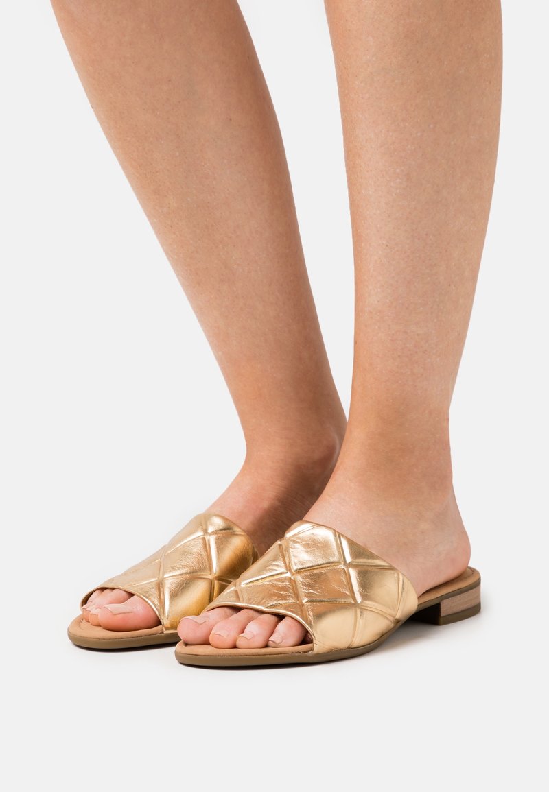 Pieds portant des sandales plates ouvertes à bout ouvert matelassées or métallisé avec un petit talon en bois sur fond blanc.