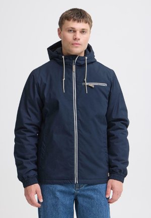 Jeune homme portant une veste à capuche bleu marine avec une fermeture éclair frontale et une poche poitrine zippée, associée à un jean bleu.