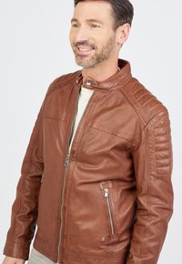 Veste de moto en cuir marron avec un design matelassé aux épaules, fermeture éclair sur le devant, deux poches zippées et un col à boutons-pression.
