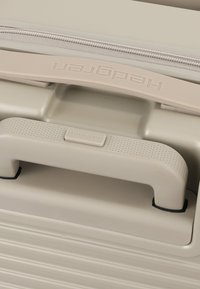 Poignée beige de valise coque dure avec prise texturée et logo Hedgren embossé sous une couture de fermeture éclair.