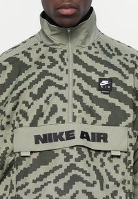 Žalias kamufliažo vėjo striukė su pusiau užtrauktuku, aukštu kaklu ir juodu "NIKE AIR" juostele per krūtinę; turi tekstūrinį raštą ir logotipo pleistrą.
