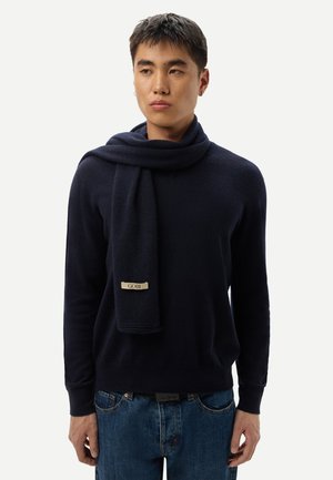 Junger Mann trägt einen marineblauen Pullover und passenden marineblauen Schal mit der Aufschrift "GOBI", kombiniert mit blauen Jeans, stehend vor einem einfarbigen hellen Hintergrund.