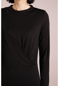 Svart långärmad topp med hög halsringning och draperad detaljerad front. Tillverkad av texturerad tyg och har en figurnära silhuett.