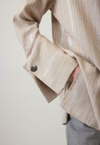 Bežina košulja sa srebrnim prugama, sa širokim rukavima i dugmetom na manžetama. Materijal izgleda gladak i lagan.