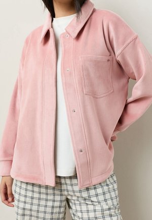 Personne portant une chemise en velours côtelé rose clair boutonnée sur un haut blanc et un pantalon à carreaux noir et blanc.