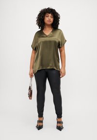 Top in satin verde oliva con scollatura a V e maniche corte; abbinato a leggings neri attillati e tacchi alti neri con cinturini, mentre tiene una pochette.