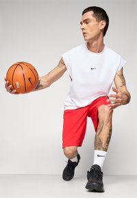 Young man in white sleeveless shirt and red shorts dribbling a basketball, wearing black sneakers and white Nike socks, against a plain background.