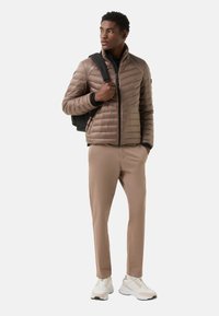 Bruine pufferjas met een doorgestikt ontwerp, gecombineerd met lichtbruine broek en witte sneakers. Model draagt een zwarte rugzak.