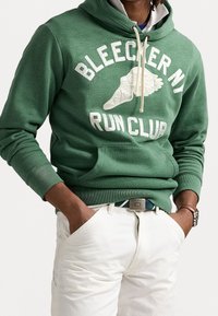 Man som bär en grön "Bleecker NY Run Club" huvtröja med krämfärgade shorts, händerna i fickorna och en klocka på vänster arm.