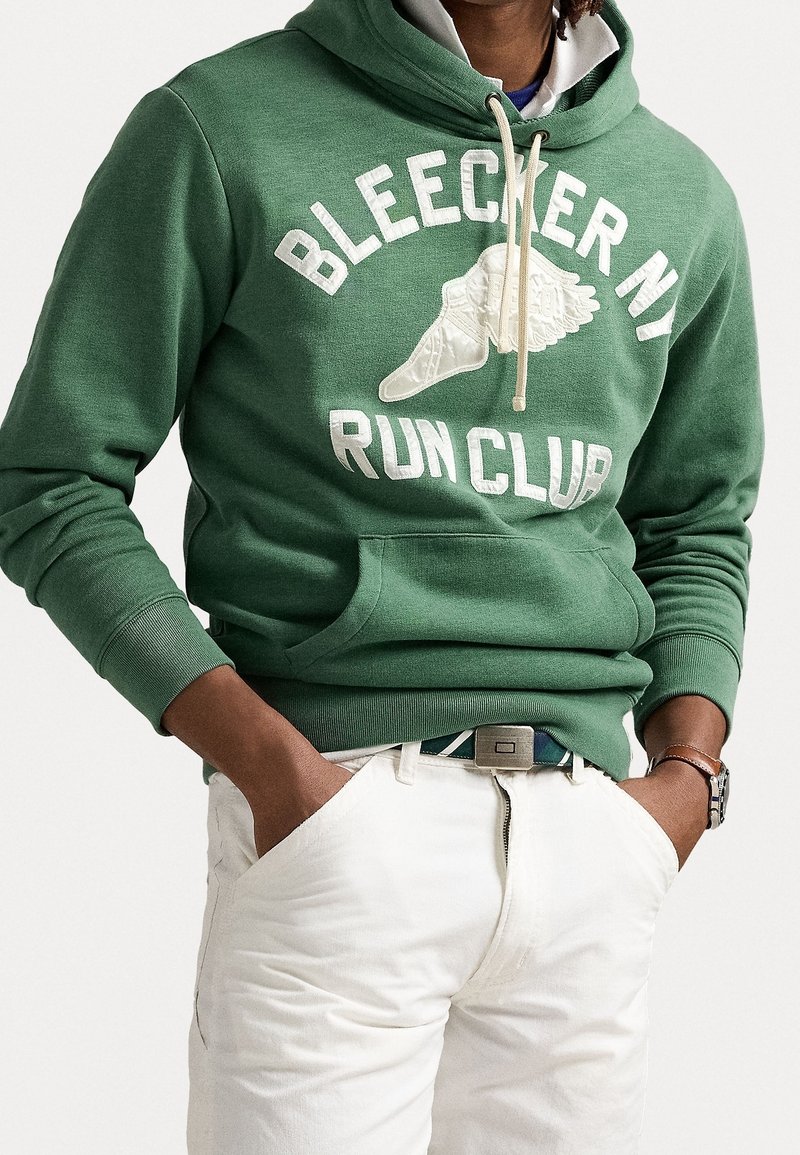 Man som bär en grön "Bleecker NY Run Club" huvtröja med krämfärgade shorts, händerna i fickorna och en klocka på vänster arm.