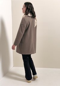 Lichtbruin jasje met een eenvoudig ontwerp, voorzien van een gladde textuur en een knoopsluiting. Gepaard met donkere flared broek en beige schoenen.