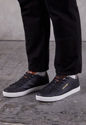 Pieds chaussés de baskets noires Jack & Jones avec semelles blanches, assorties à un pantalon noir, debout sur un sol gris texturé.