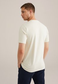 Tricou scurt, de culoare bej deschis, fabricat dintr-un material neted, cu guler rotund, croi lejer și o textură subtilă pe spate.