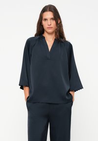 Dunkelblaue Bluse mit V-Ausschnitt und 3/4-Ärmeln, die über gezackte Kanten verfügt. Hergestellt aus einem glatten, fließenden Material mit lockerer Passform.