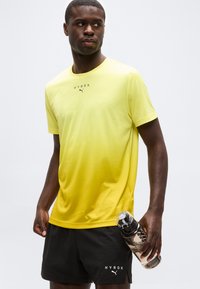 Άνδρας που φοράει κίτρινο T-shirt HYROX Puma και μαύρο σορτς, κρατώντας ένα μπουκάλι νερό, κοιτάζει προς τα αριστερά του σε απλό φόντο.