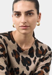 Beżowy sweter w czarno-brązowy wzór w panterkę, z okrągłym dekoltem. Złote kolczyki w kształcie kół dodają metalicznego akcentu. Gładka tekstura.