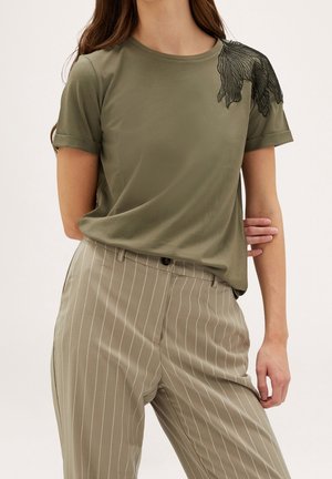 Camiseta de manga corta verde oliva con un diseño texturizado de hojas negras en el hombro, combinada con pantalones gris claro a rayas.