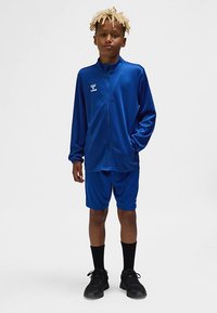 Blauw sporttrainingspak van lichtgewicht stof, bestaande uit een jasje met rits en bijpassende korte broek. Voorzien van logodetail en zwarte schoenen.