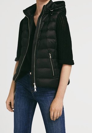 Person trägt schwarze gesteppte ärmellose Jacke mit Reißverschluss über schwarzem Stricktop und blauen Jeans, steht vor einfarbigem Hintergrund.