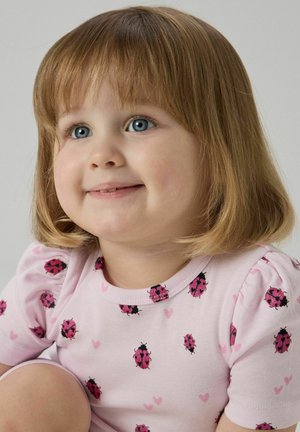 Maluch z jasnobrązowym bobem, niebieskimi oczami, ubrany w różową koszulkę z wzorami biedronek i serduszek, uśmiechający się i patrzący lekko w górę.