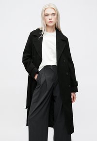 Svart ull trenchcoat med store skjorteklaffer, dobbelknappede knapper og sidelommer, paret med grå høyt livbukser.