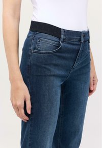 Blugi din denim de culoare albastru închis, cu un brâu elastic negru, având cinci buzunare și un design cu picior drept, prezentate din lateral.