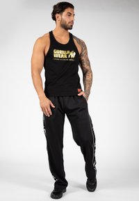 Schwarzes Tanktop mit goldener "GORILLA WEAR" Schrift, kombiniert mit schwarzen Hosen mit weißen seitlichen Streifen. Das Model trägt schwarze Sneakers.