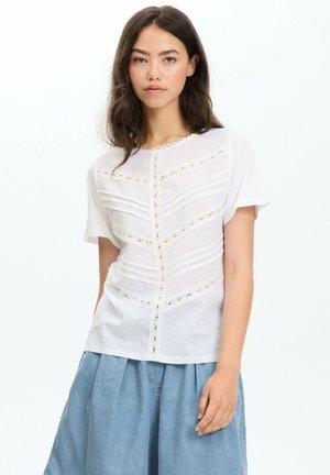 Joven mujer con cabello largo y oscuro lleva una blusa blanca con detalles de encaje y una falda de mezclilla azul suelta, de pie frente a un fondo blanco liso.