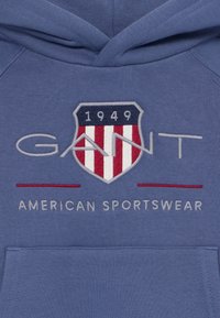 Modrá bavlnená mikina s kapucňou so štítovým logom s textom "1949", červenými a bielymi pruhmi a vyšitým nápisom "GANT AMERICAN SPORTSWEAR" pod ním.