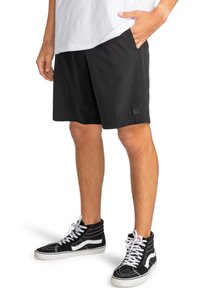 Pantalones cortos negros hechos de un tejido ligero, con un corte relajado, bolsillos laterales y una pequeña etiqueta de marca en el dobladillo. Combinados con zapatillas negras.