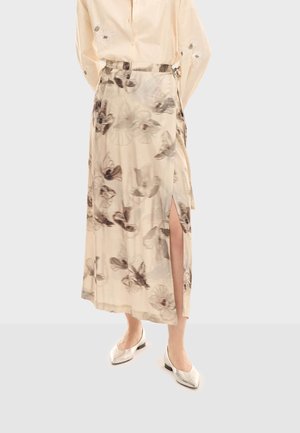 Žena nosi bež midi suknju s cvjetnim uzorkom i prorezom na bedrima, krem košulju s izvezenim rukavima i srebrne šiljaste balerinke.