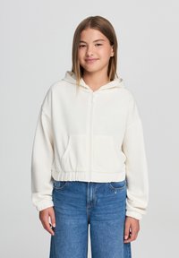 Felpa corta con zip in pile bianco, dotata di cappuccio, polsini a costine e tasca frontale, abbinata a jeans blu a vita alta.