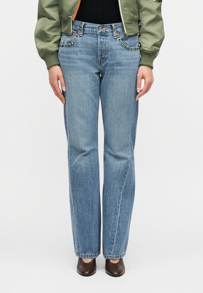 Γυναίκα που φοράει ανοιχτό μπλε τζιν με ίσια μπατζάκια και διακοσμητικές τρουκς στα τσέπες, καθώς και καφέ παπούτσια με τετράγωνη μύτη, σετ με πράσινο cropped σακάκι.
