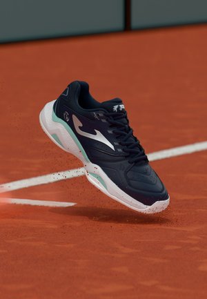 Mustvalge tennisesaabas keset õhku saviliivalisel väljakul, punaste saviosakestega tallal ümbritsetud.