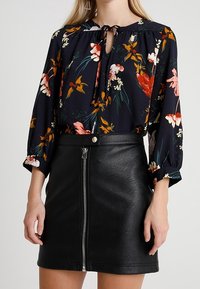 Bloemige blouse met lange mouwen en een strikdetail bij de hals, gecombineerd met een zwarte leren minirok met hoge taille en een ritssluiting aan de voorkant.